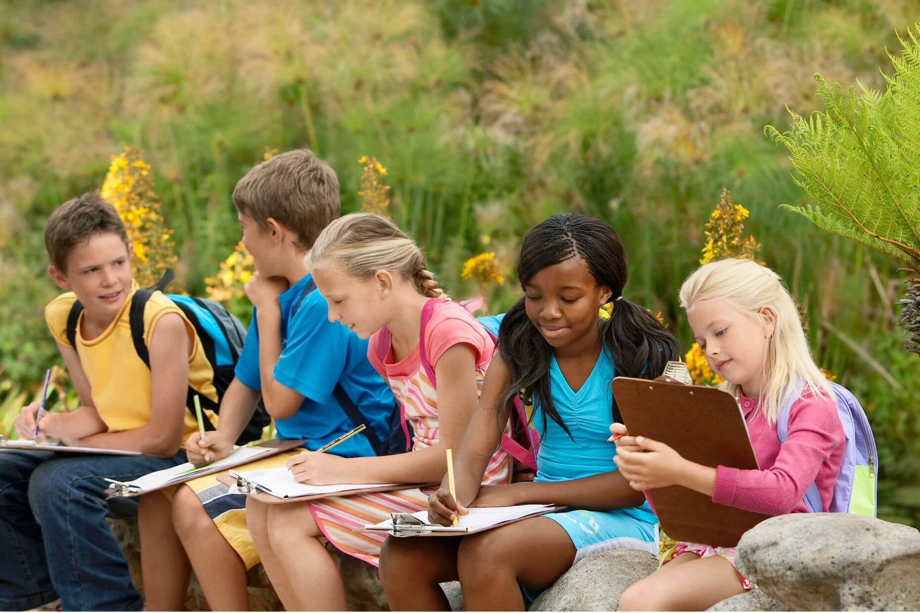 Schulkinder sitzen vor Blumen im Freien und schreiben.