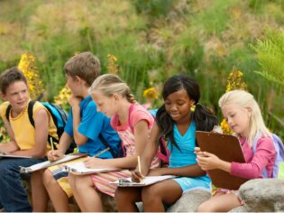 Schulkinder sitzen vor Blumen im Freien und schreiben.